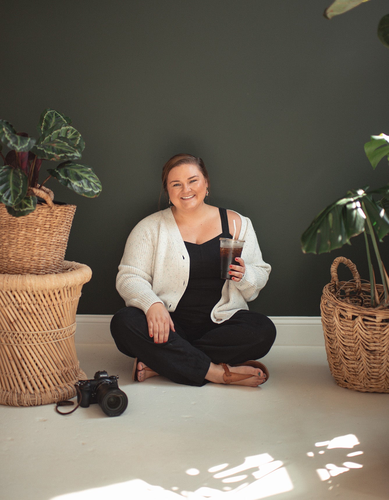 Person sitting on the floor with plants and a camera in a room with a dark wall and light floor.