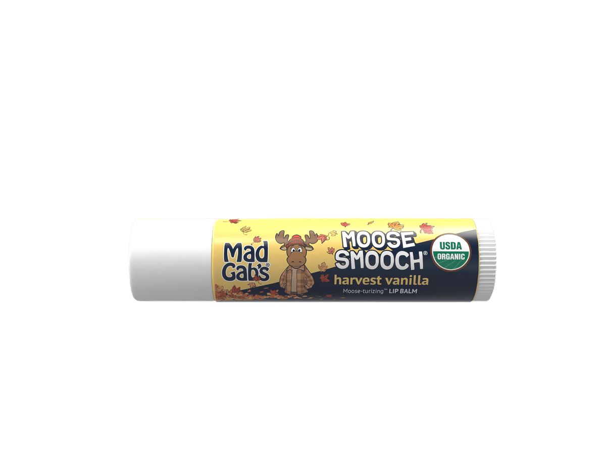 A tube of Moose Smooch lip balm by Mad Cats, in Harvest Vanilla flavor. The packaging features a yellow and dark blue label with a cartoon moose wearing a plaid shirt and autumn leaves, alongside a green USDA Organic seal.